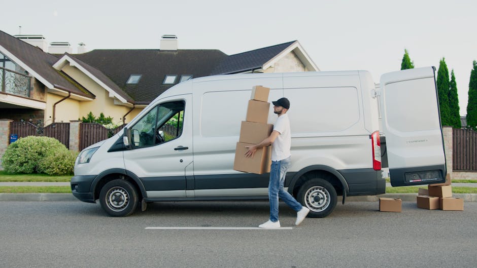 A man dressed in casual clothing, including a white T-shirt, blue jeans, white sneakers, and a black cap, is carrying three cardboard boxes of varying sizes in front of a white panel van parked on the