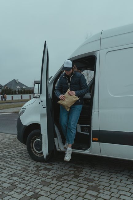 A man dressed in casual clothing, including a white T-shirt, blue jeans, white sneakers, and a black cap, is carrying three cardboard boxes of varying sizes in front of a white panel van parked on the