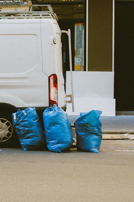 A white delivery van parked outside a building with a glass door, showing its rear doors partially open. In front of the van, three large blue plastic rubbish bags filled with packing materials or unw