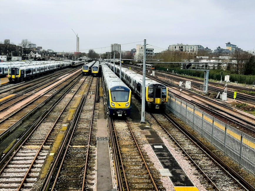 A view of multiple modern trains parked on railway tracks at a station, with the closest trains in focus featuring yellow and grey livery and positioned side by side, while additional train carriages extend further into the distance on parallel tracks. The platform edges are visible on the right, with a metal fence separating them from the tracks. Overhead electrical wires and supporting poles stretch across the scene, indicating a well-organised rail infrastructure. In the background, there are several buildings, including mid-rise and high-rise structures, and some green spaces, under an overcast sky. The scene captures an urban transportation hub that could be associated with the logistics of house removals or furniture transport, as part of moving or relocation services provided by companies like Man and Van Pinner, which specialise in efficient home relocation and furniture transport within the UK.