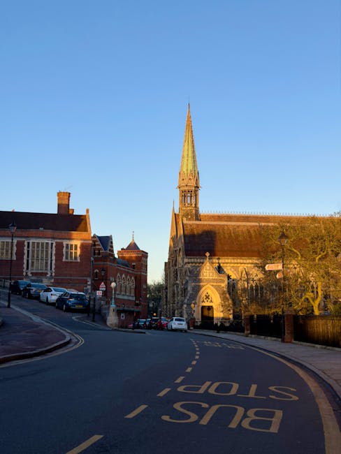 An outdoor street scene during daylight showing a historic church with a tall, pointed spire and Gothic architectural features, situated beside a row of red-brick buildings. The street has a curved road marked with yellow 'BUS STOP' lines and curbside parking filled with various cars. In the foreground, the road signage and pavement are visible, with the church's stone facade and arched entrance partly shaded by nearby trees. This setting illustrates an urban environment where house removals and furniture transport may take place, highlighting the importance of careful planning during home relocations. Man and Van Pinner offers professional removals services, including loading, packing, and transport, seamlessly integrated into such busy street environments.