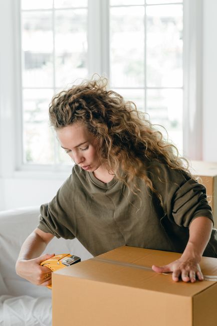 A woman with curly, shoulder-length hair wearing a loose-fitting olive green top is indoors, preparing a cardboard box for packing. She is using a tape dispenser to seal the box, which is positioned o