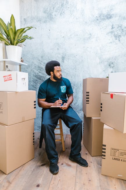 A woman with curly, shoulder-length hair wearing a loose-fitting olive green top is indoors, preparing a cardboard box for packing. She is using a tape dispenser to seal the box, which is positioned o
